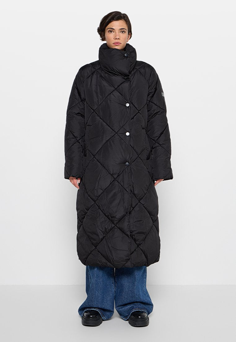 Schwarzer langer Steppmantel mit hohem Kragen, diamantförmigem Steppmuster und Druckknöpfen; getragen über weit geschnittene blaue Jeans und schwarzen Schuhen.