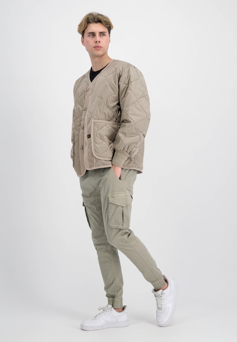 Beigefarbene gestepptes Jacke mit V-Ausschnitt, vorderen Knöpfen und Seitentaschen, kombiniert mit hellgrünen Cargo-Hosen und weißen Sneakers.