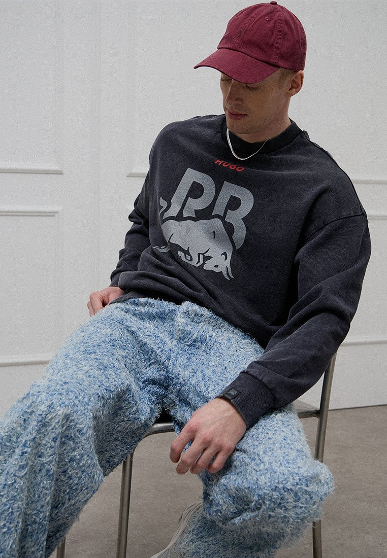 Schwarzer Sweatshirt mit grauem Grafikdruck und roten Akzenten, kombiniert mit flauschigen, blau strukturierten Hosen. Modell trägt eine bordeauxrote Baseballkappe.
