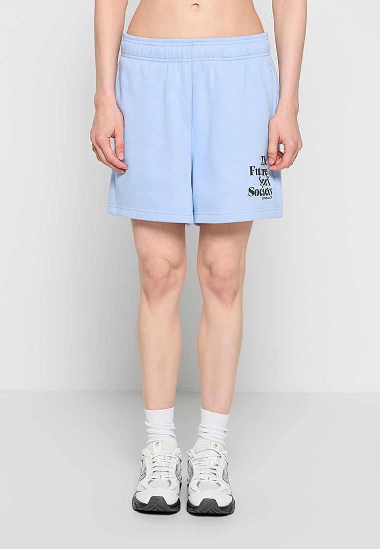Person trägt hellblaue Shorts mit schwarzem Text am linken Bein, weiße Socken und weiße Sportschuhe, steht vor einem schlichten Hintergrund.