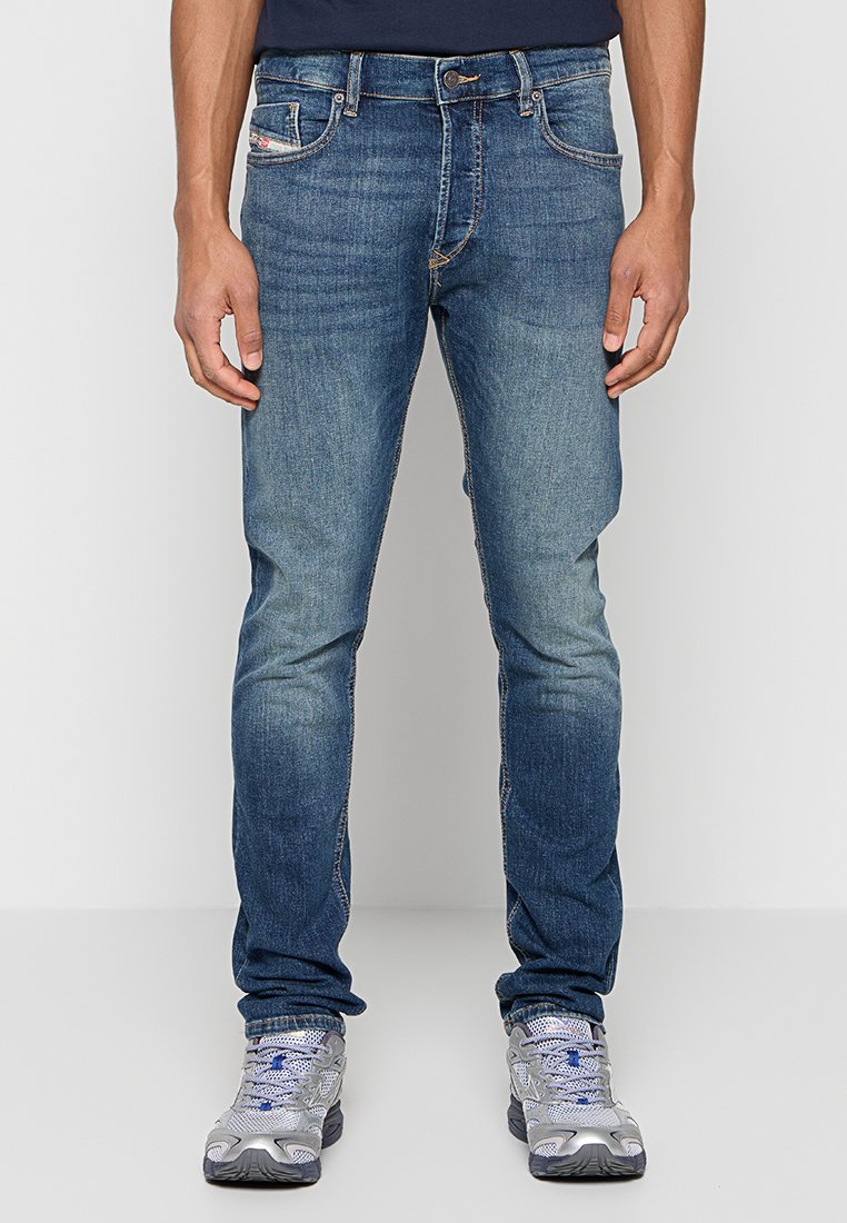 Schmal geschnittene Jeans aus Denim in mittlerer blauer Waschung mit verblassten Akzenten. Verfügt über fünf Taschen, Gürtelschlaufen und kontrastierende Nähte. Kombiniert mit silbernen Sportschuhen.