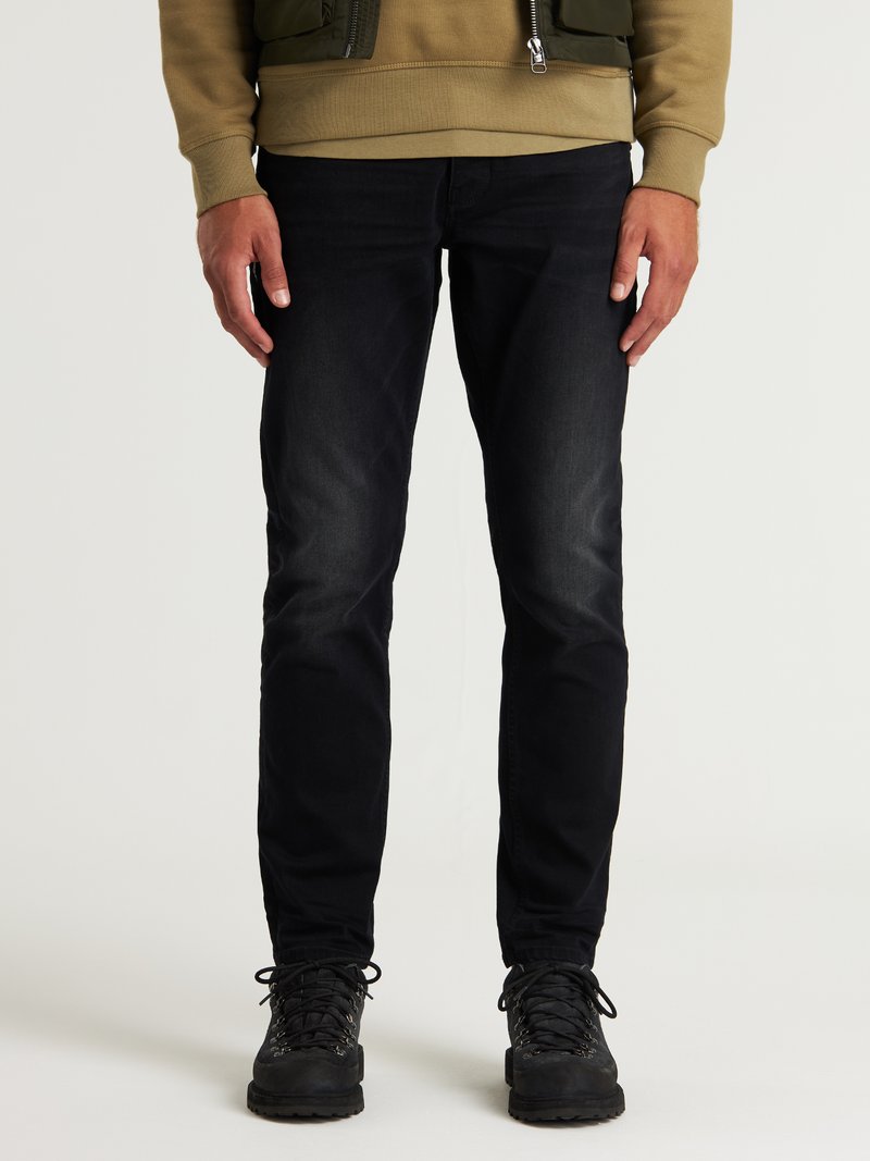 Schwarze Jeans aus Denim mit Slim-Fit, kombiniert mit einem hellbraunen Sweatshirt. Das Outfit umfasst robuste schwarze Wanderstiefel und dezente Texturdetails.