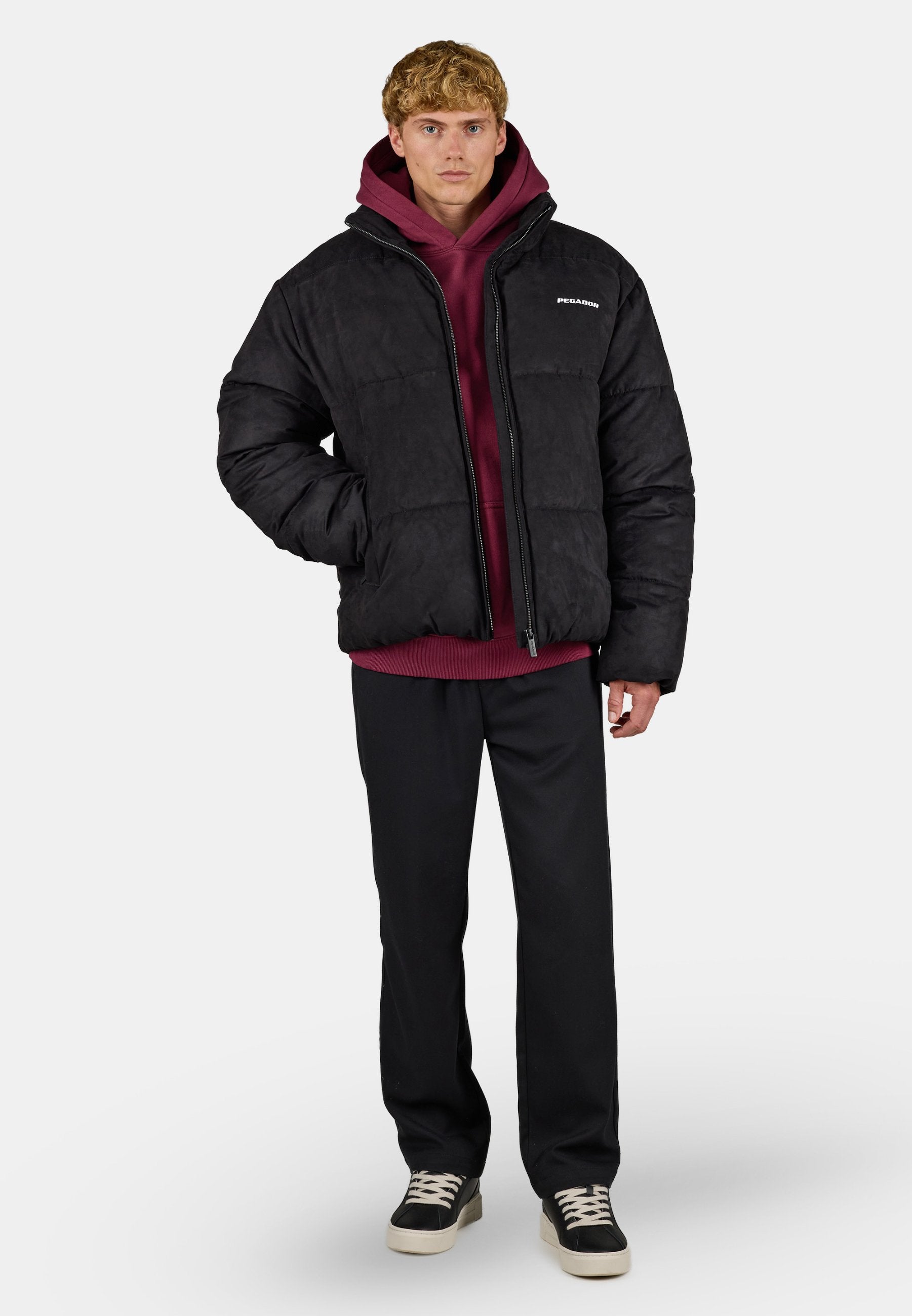 Junger Mann trägt eine schwarze Steppjacke über einem bordeauxfarbenen Hoodie, schwarze Hose und schwarze Sneaker mit weißen Sohlen und Schnürsenkeln.