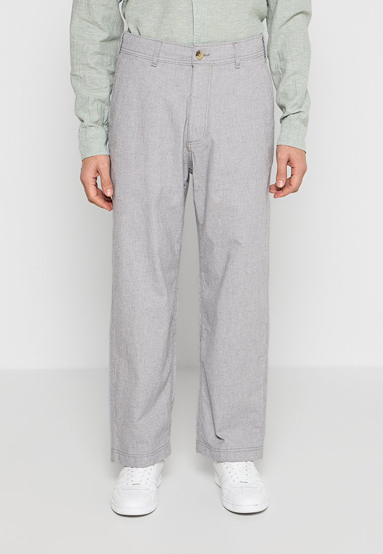 Mann trägt hellgraue, gerade geschnittene Hose, hellgrünes Langarmhemd und weiße Sneakers, steht vor einem einfarbigen Hintergrund.