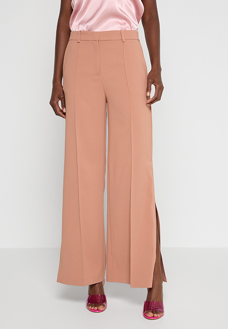 Pfirsichfarbene weit geschnittene Hose aus weichem Stoff mit seitlichen Schlitzen; verfügt über eine hohe Taille und FaltenDetails, ergänzt durch glänzende pinkfarbene Schuhe.