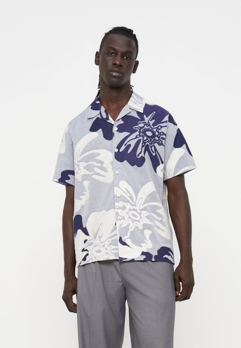 Mann trägt kurzärmliges Hemd mit großem weiß-blauem Blumenmuster und graue Hose vor schlichtem Hintergrund.