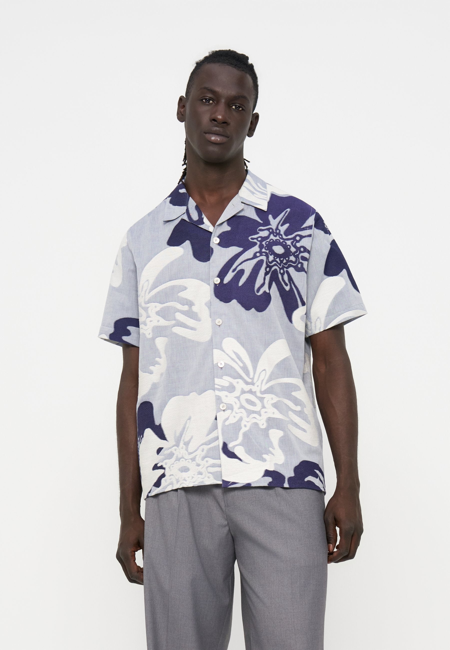 Mann trägt kurzärmliges Hemd mit großem weiß-blauem Blumenmuster und graue Hose vor schlichtem Hintergrund.