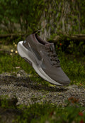 Brauner und weißer Nike Trailrunning-Schuh mit Gore-Tex, in der Luft über einem Kiesweg im Wald.