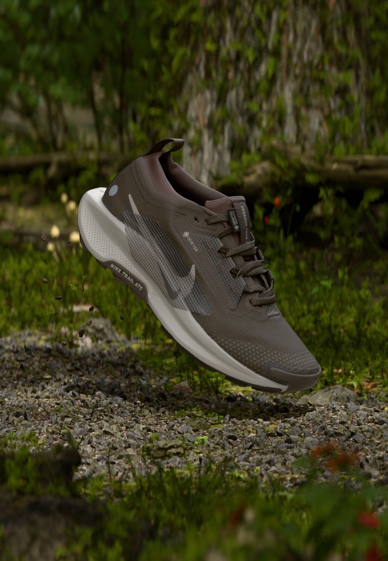 Brauner und weißer Nike Trailrunning-Schuh mit Gore-Tex, in der Luft über einem Kiesweg im Wald.