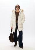 Weißer Puffer-Mantel mit einer Kapuze aus Pelzbesatz, kombiniert mit einem braunen Oberteil und dunkelblauen, ausgestellten Jeans, hält eine braune Wildleder-Handtasche. Große Sonnenbrille.