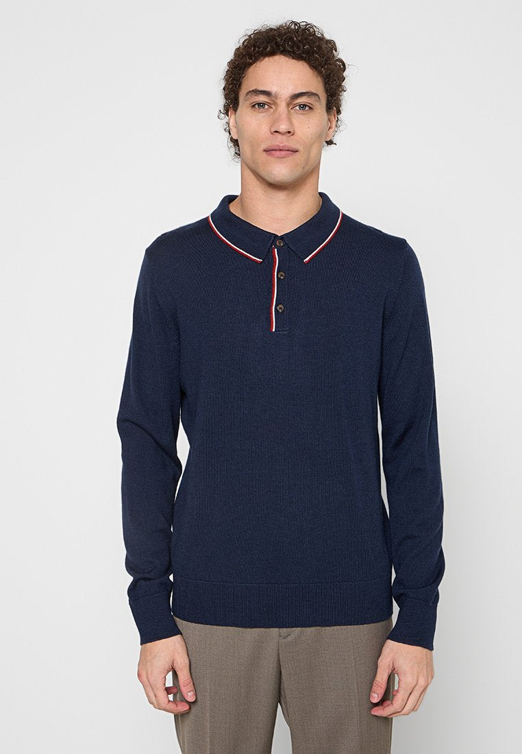 Mann mit lockigem Haar, der ein navyblaues Langarm-Polohemd mit rot-weißen Akzenten am Kragen und braunen Hosen trägt, steht vor einem einfarbigen Hintergrund.