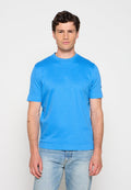 Mann steht frontal, trägt ein leuchtend blaues Kurzarmhemd und hellblaue Jeans vor einem schlichten, hellen Hintergrund.