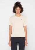 Person mit lockigem Haar trägt ein schlichtes beige Kurzarmshirt und dunkle Hosen und steht vor einem einfarbigen weißen Hintergrund.