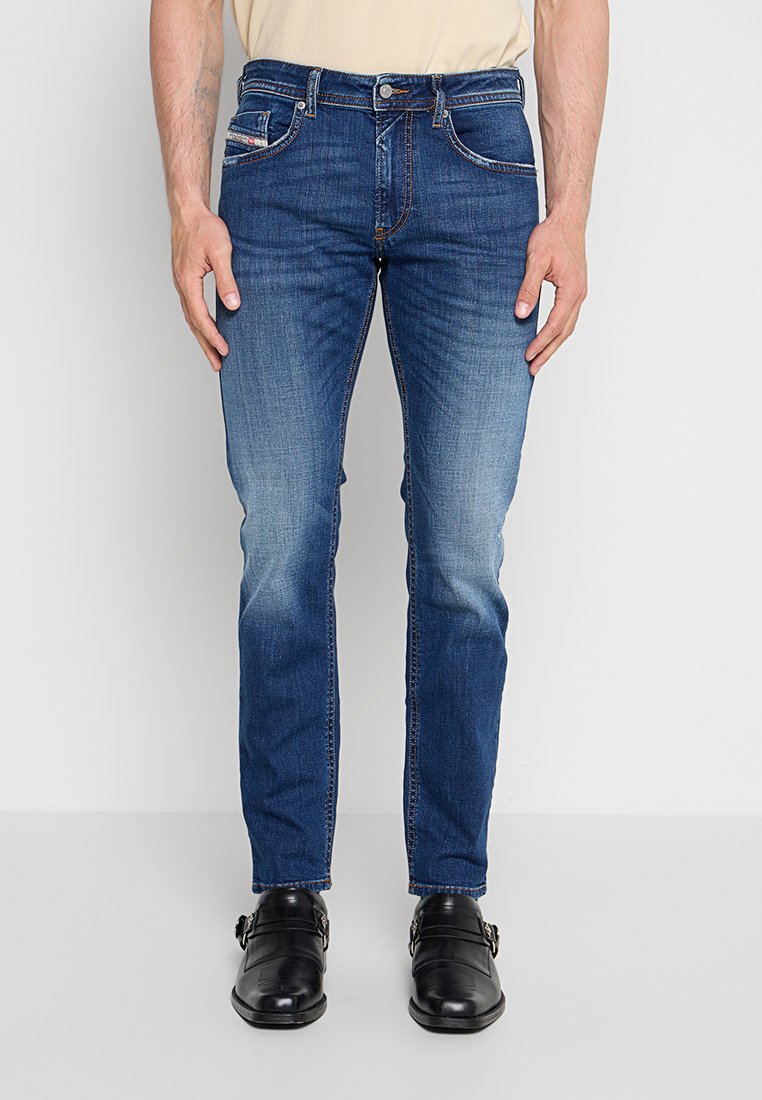 Mann trägt Slim-Fit blaue Denim-Jeans und schwarze Leder Monk-Strap Schuhe, stehend vor einem schlichten hellgrauen Hintergrund.