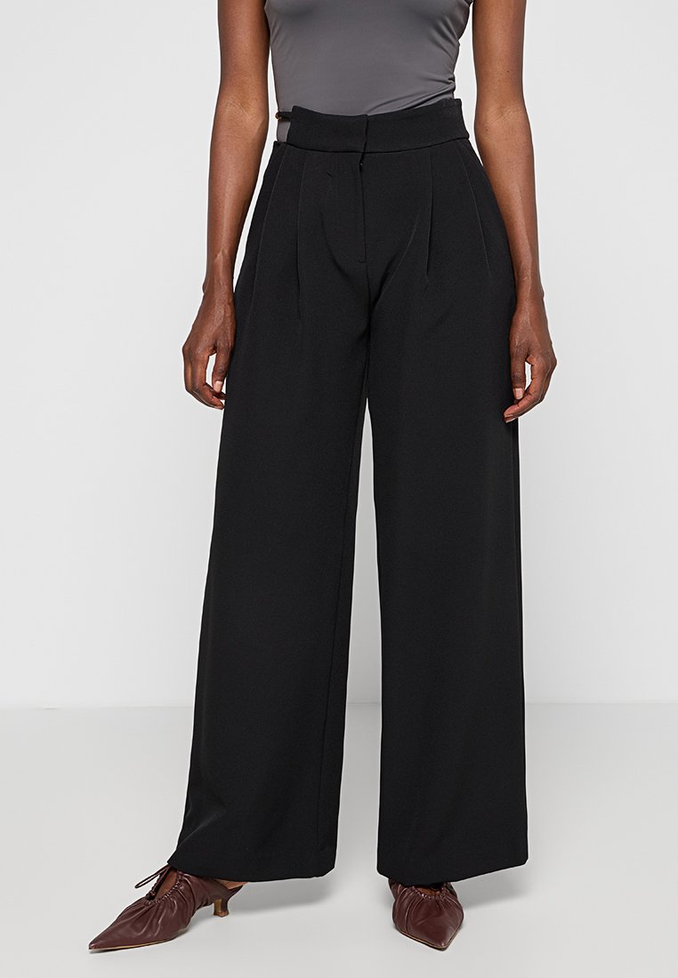 Weite schwarze Hose mit hoher Taille, gefaltetem Vorderteil und glatter Stofftextur. Gestylt mit einem figurbetonten grauen Oberteil und braunen Schuhen.