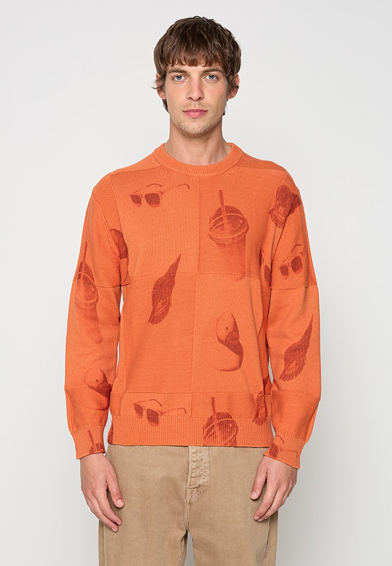 Mann trägt orangefarbenen Pullover mit aufgedruckten Motiven von Sonnenbrille, Getränk mit Strohhalm, Feder und Vogelkopf, kombiniert mit beiger Hose.