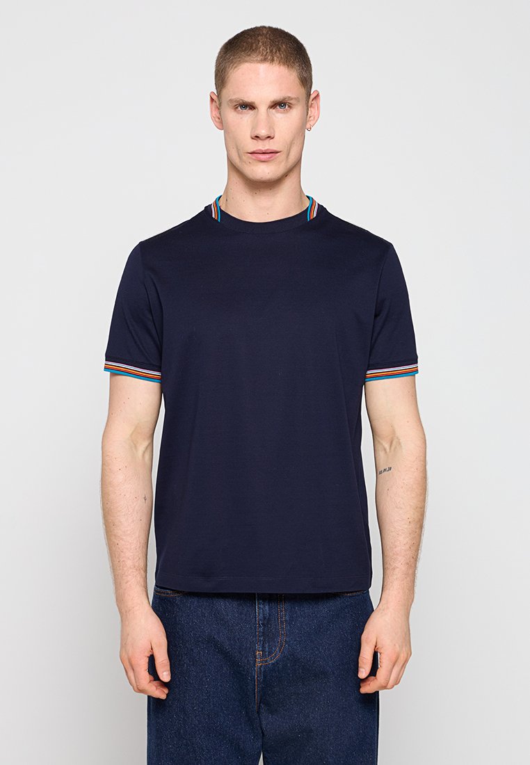 Junger Mann mit kurzen Haaren, der ein marineblaues T-Shirt mit mehrfarbig gestreiftem Besatz an Ärmeln und Kragen trägt, kombiniert mit dunkelblauen Jeans.