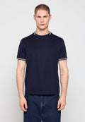 Junger Mann mit kurzen Haaren, der ein marineblaues T-Shirt mit mehrfarbig gestreiftem Besatz an Ärmeln und Kragen trägt, kombiniert mit dunkelblauen Jeans.
