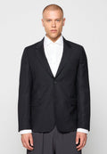 Mann in schwarzem Ein-Knopf-Blazer über weißem Hemd und grauen Hosen, stehend vor einem einfachen hellen Hintergrund.