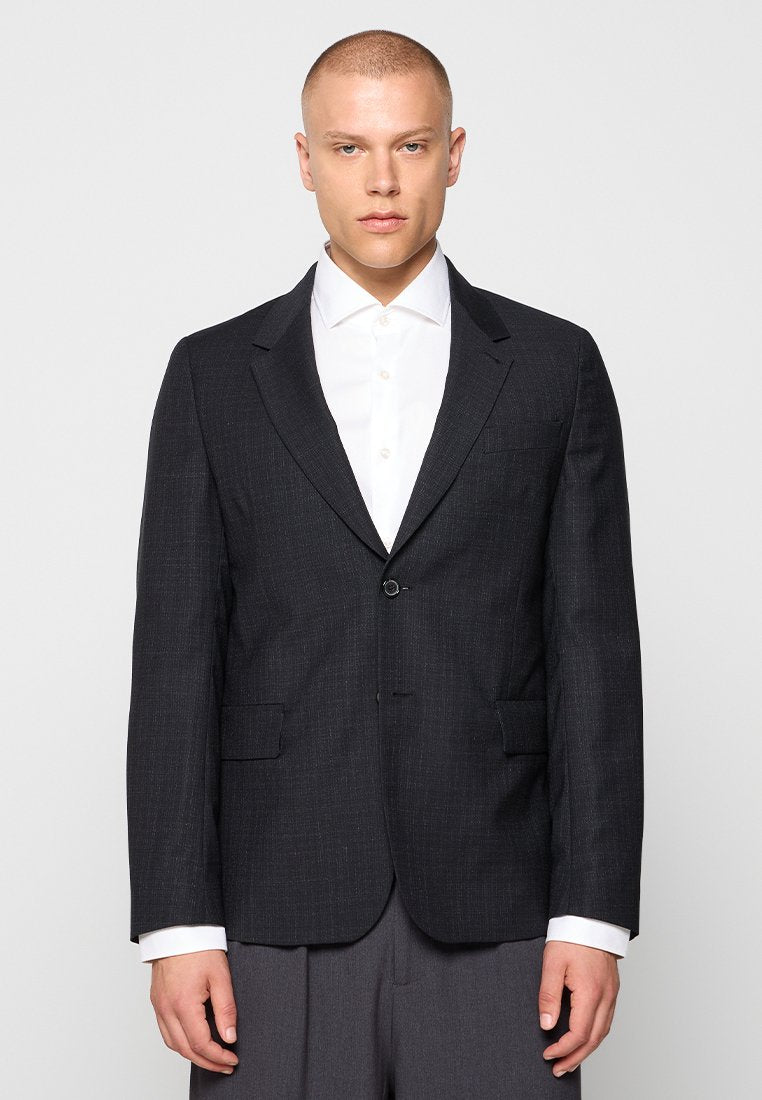 Mann in schwarzem Ein-Knopf-Blazer über weißem Hemd und grauen Hosen, stehend vor einem einfachen hellen Hintergrund.