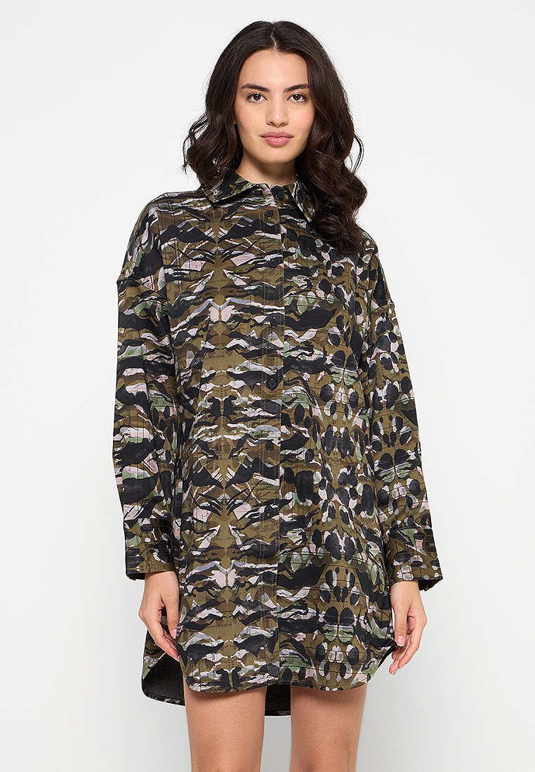Junge Frau mit langen, dunklen Haaren, die ein langärmeliges, knöpfbares Hemdkleid im Camouflage-Muster trägt und vor einem schlichten Hintergrund steht.