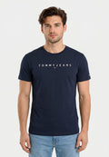 Junger Mann mit kurzen braunen Haaren trägt ein marineblaues Tommy Jeans T-Shirt und hellblaue Jeans, steht vor einem schlichten weißen Hintergrund.