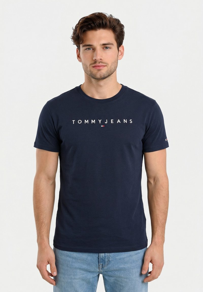 Junger Mann mit kurzen braunen Haaren trägt ein marineblaues Tommy Jeans T-Shirt und hellblaue Jeans, steht vor einem schlichten weißen Hintergrund.