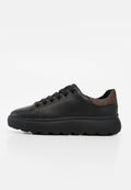 Schwarzer Low-Top-Sneaker mit dicker Sohle, schwarzen Schnürsenkeln und braunem strukturiertem Fersenpatch, von der Außenseite auf weißem Hintergrund gezeigt.
