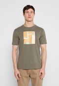 Mann trägt ein olivgrünes T-Shirt mit beige-weißem geometrischem Quadratmuster, kombiniert mit hellbraunen Hosen, steht vor einem einfarbigen Hintergrund.