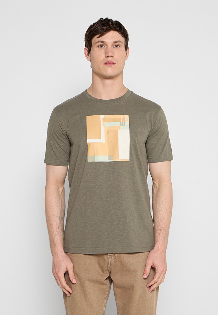 Mann trägt ein olivgrünes T-Shirt mit beige-weißem geometrischem Quadratmuster, kombiniert mit hellbraunen Hosen, steht vor einem einfarbigen Hintergrund.