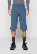 Mann trägt blaue knielange Shorts, graues langärmeliges Oberteil, graue Socken und schwarze Sportschuhe, steht vor einem schlichten Hintergrund.