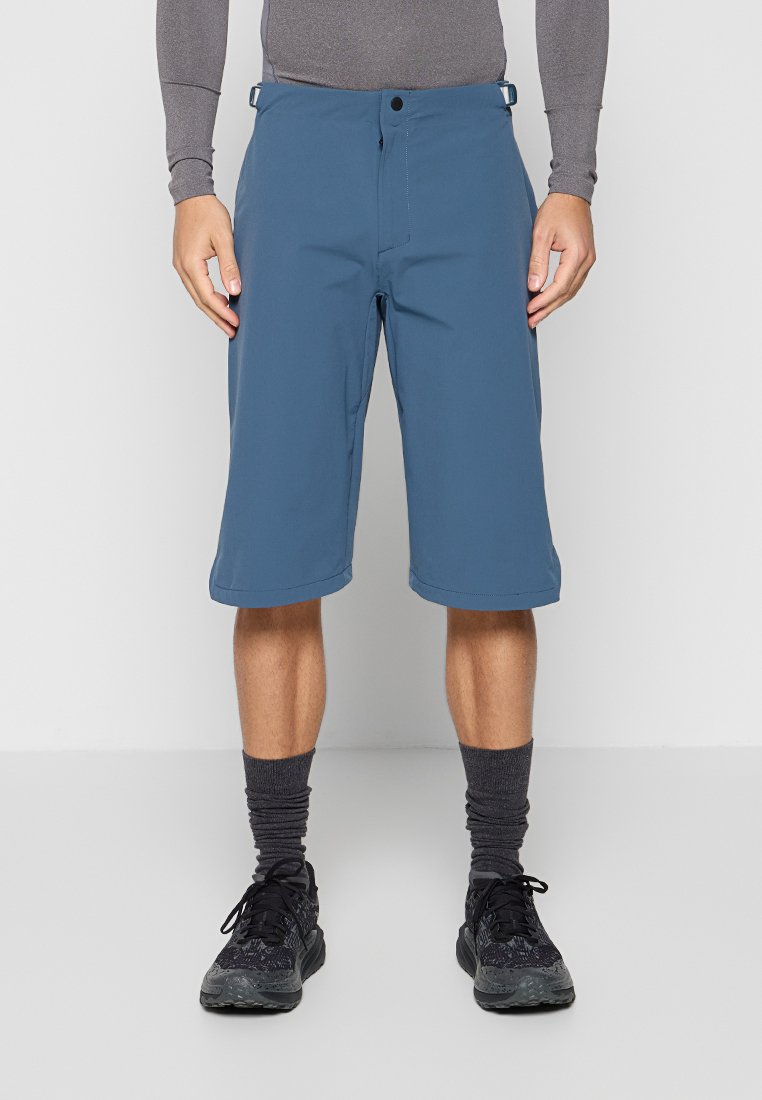Mann trägt blaue knielange Shorts, graues langärmeliges Oberteil, graue Socken und schwarze Sportschuhe, steht vor einem schlichten Hintergrund.