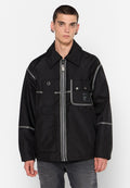 Junger Mann in einer schwarzen Jacke mit Reißverschluss, die mehrere Taschen und reflektierende Streifen hat, kombiniert mit hellgrauen Jeans, stehend vor einem weißen Hintergrund.