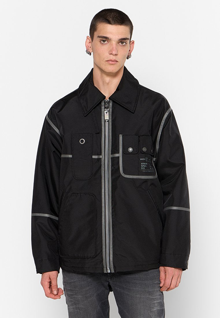 Junger Mann in einer schwarzen Jacke mit Reißverschluss, die mehrere Taschen und reflektierende Streifen hat, kombiniert mit hellgrauen Jeans, stehend vor einem weißen Hintergrund.