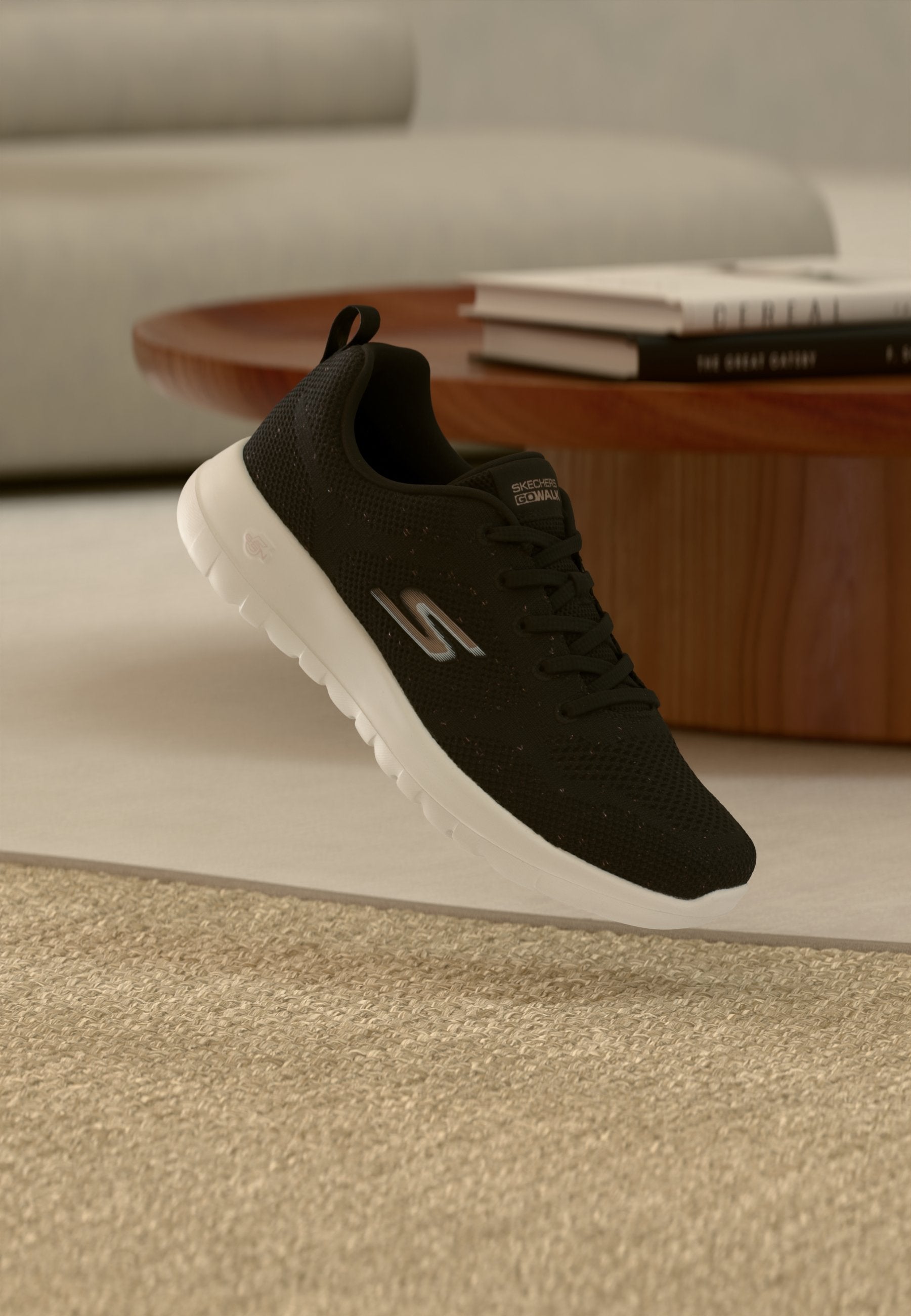 Schwarzer Skechers-Sneaker mit weißer Sohle, der über einem beigen Teppich schwebt, im Hintergrund runder Holztisch und gestapelte Bücher.