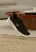 Schwarzer Skechers-Sneaker mit weißer Sohle, der über einem beigen Teppich schwebt, im Hintergrund runder Holztisch und gestapelte Bücher.