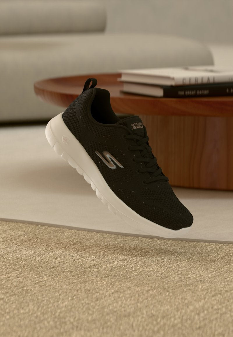Schwarzer Skechers-Sneaker mit weißer Sohle, der über einem beigen Teppich schwebt, im Hintergrund runder Holztisch und gestapelte Bücher.