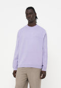 Mann mit dunkler Haut und geflochtenem Haar, der einen hellvioletten Pullover und beige Hose vor weißem Hintergrund trägt.