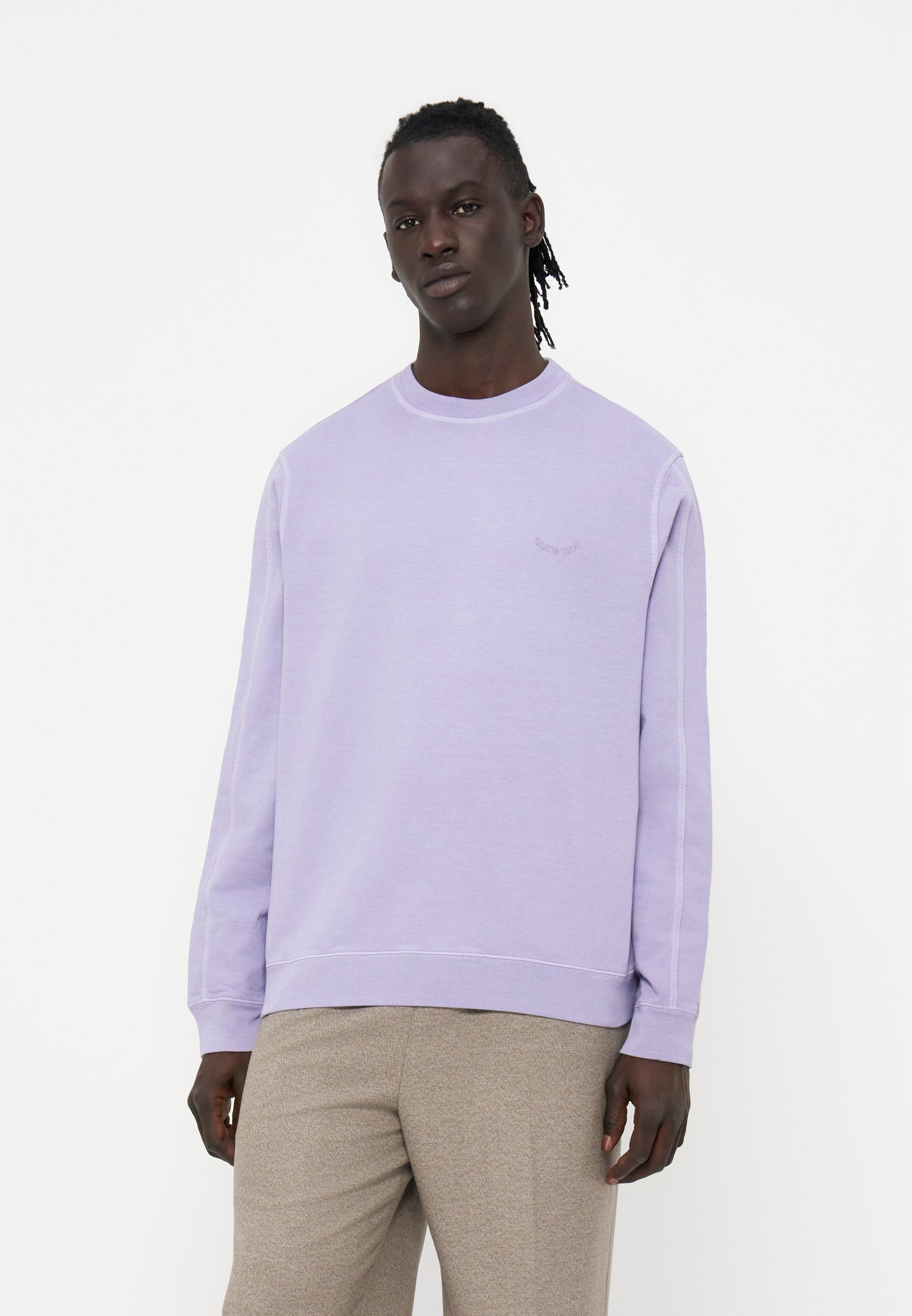 Mann mit dunkler Haut und geflochtenem Haar, der einen hellvioletten Pullover und beige Hose vor weißem Hintergrund trägt.