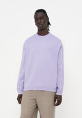 Mann mit dunkler Haut und geflochtenem Haar, der einen hellvioletten Pullover und beige Hose vor weißem Hintergrund trägt.