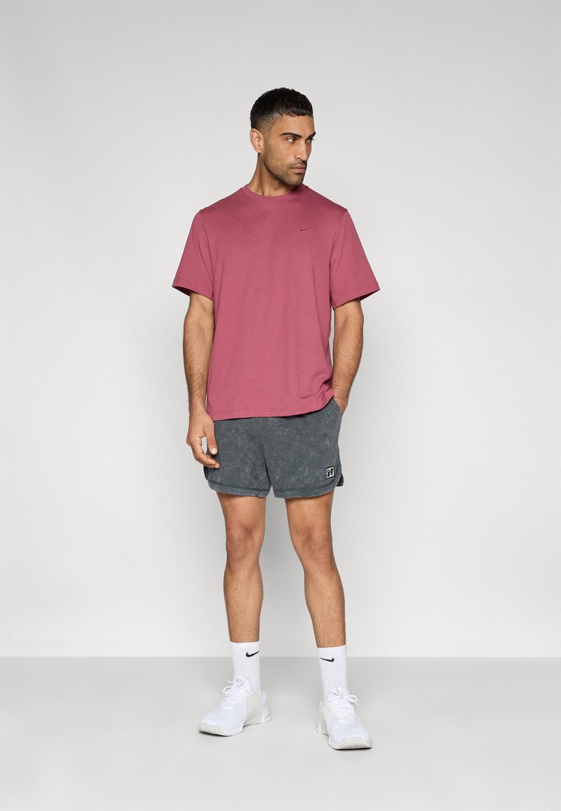Herren-T-Shirt mit kurzen Ärmeln in Kastanienbraun von Nike, kombiniert mit dunkelgrauen, strukturierten Shorts mit Logo-Patch, weißen Socken und weißen Turnschuhen.