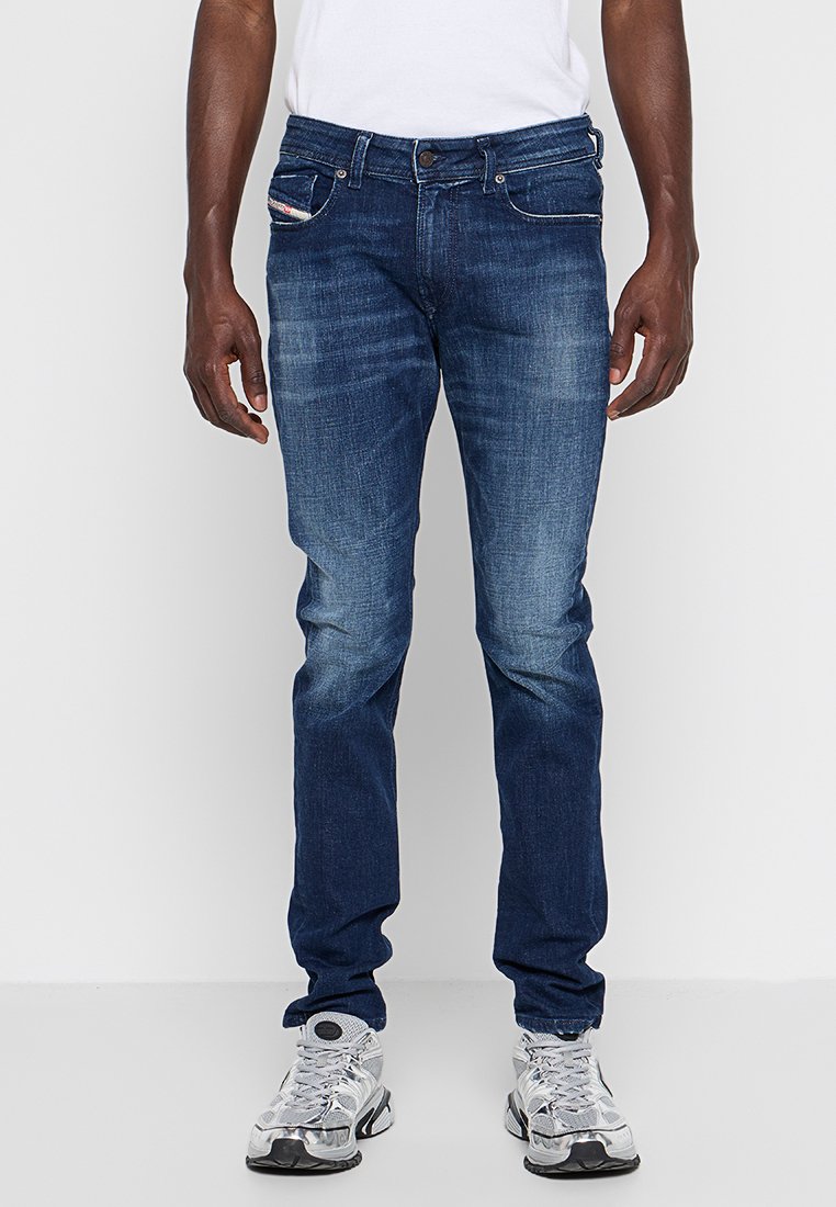 Mann in schmal geschnittenen dunkelblauen Jeans mit dezentem Verblassen und weißen Sportschuhen, der vor einem schlichten hellen Hintergrund steht.