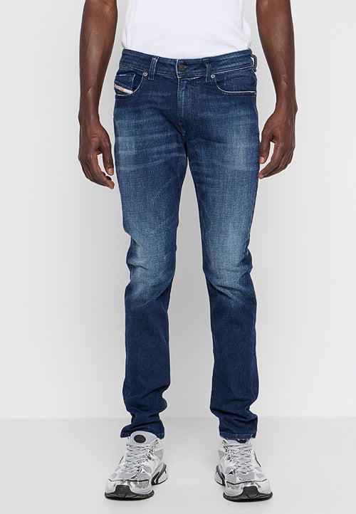 Mann in schmal geschnittenen dunkelblauen Jeans mit dezentem Verblassen und weißen Sportschuhen, der vor einem schlichten hellen Hintergrund steht.
