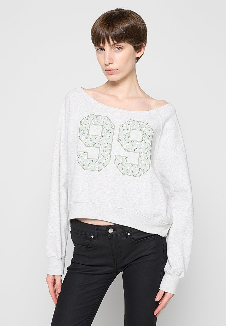 Junge Frau mit kurzen braunen Haaren, die einen hellgrauen Pullover mit der Aufschrift "99" auf der Vorderseite und schwarze, eng anliegende Hosen trägt, steht vor einem einfarbigen Hintergrund.