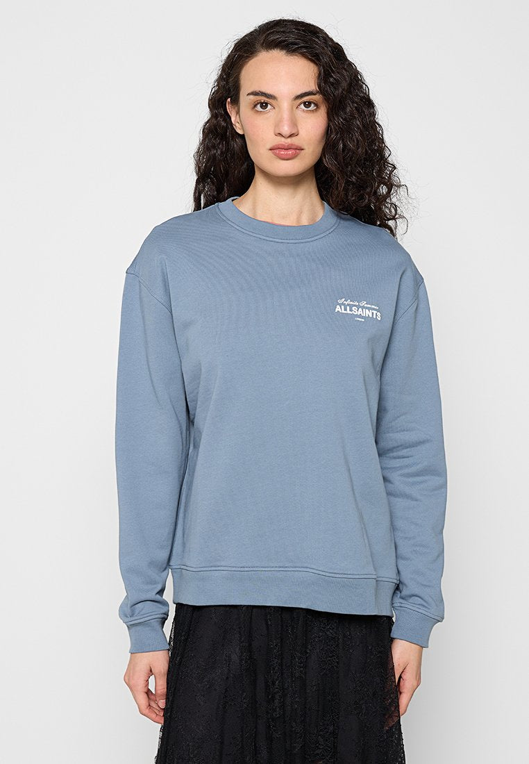 Frau mit langen, lockigen dunklen Haaren, die einen hellblauen AllSaints-Sweatshirt und einen schwarzen Spitzenrock vor einem einfarbigen hellen Hintergrund trägt.