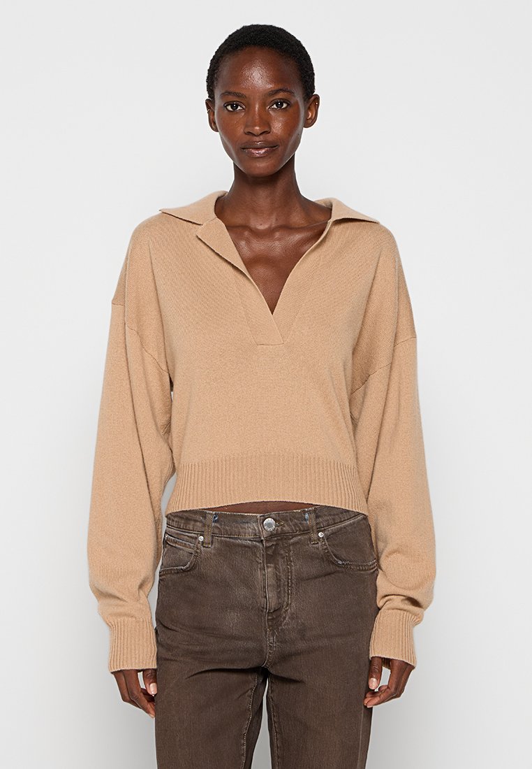 Beiger, verkürzter Pullover mit V-Ausschnitt und Kragen. Mit lockeren Ärmeln und geripptem Saum. Trägt dazu eine dunkelbraune, gerade Jeans.