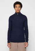 Ein Mann trägt einen dunkelblauen gemusterten Reißverschluss-Pullover mit hohem Kragen und beigen Hosen, während er vor einem schlichten hellen Hintergrund steht.