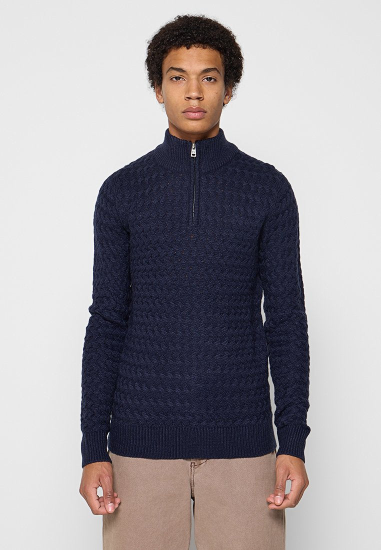Ein Mann trägt einen dunkelblauen gemusterten Reißverschluss-Pullover mit hohem Kragen und beigen Hosen, während er vor einem schlichten hellen Hintergrund steht.