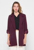 Junge Frau mit hellbraunem Haar, die einen dunkelvioletten Blazer über einem schwarzen Oberteil und weißen hochgeschnittenen Hosen trägt, vor einem weißen Hintergrund stehend.