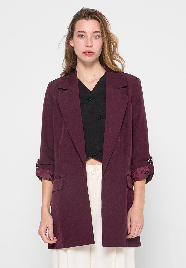 Junge Frau mit hellbraunem Haar, die einen dunkelvioletten Blazer über einem schwarzen Oberteil und weißen hochgeschnittenen Hosen trägt, vor einem weißen Hintergrund stehend.